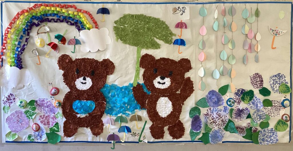 壁面飾り。熊が字葉っぱの傘をさして雨の中を散歩している様子を描いています。背景に葉自裁の花や虹が飾られています。